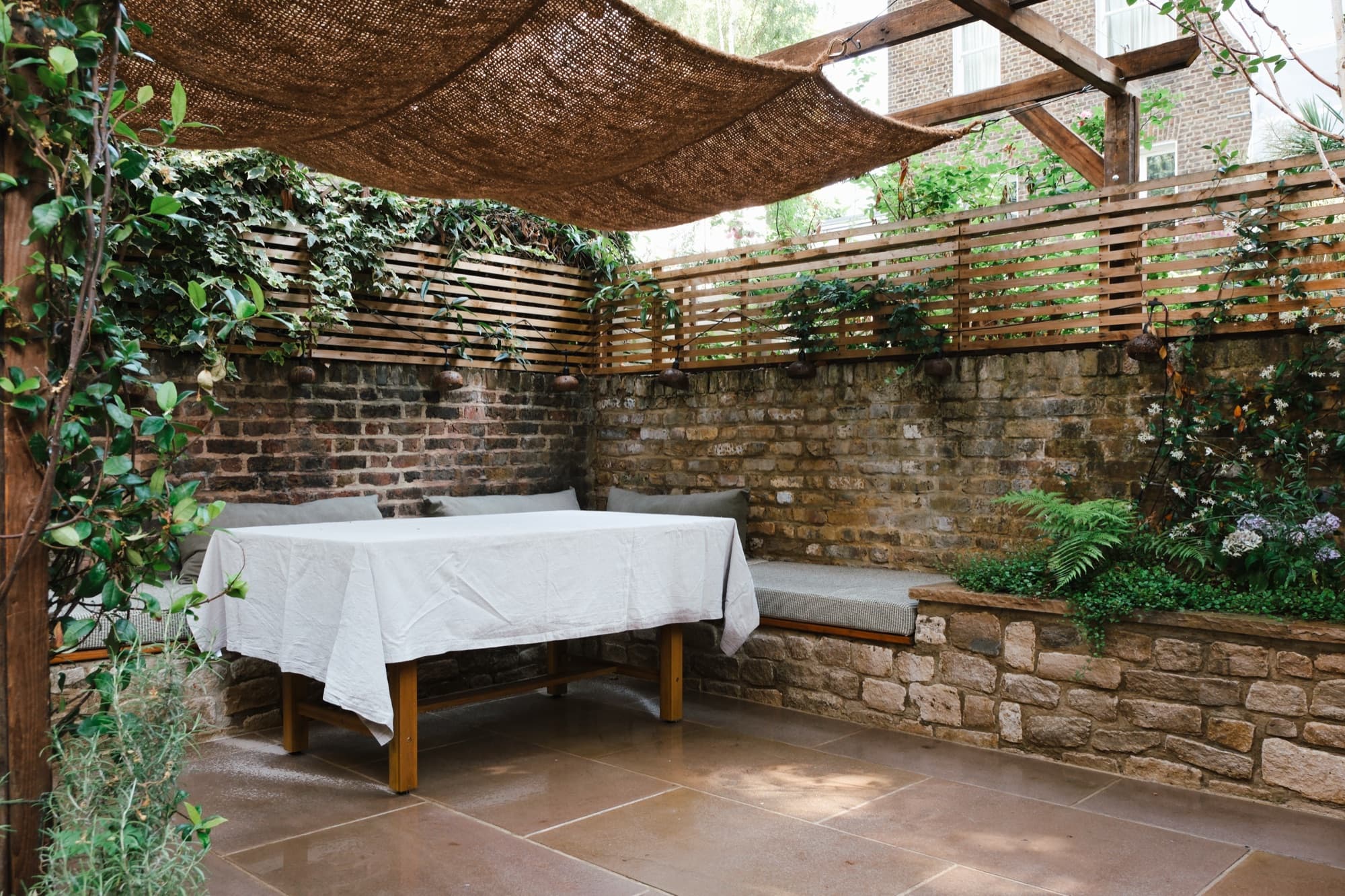 Dining table beneath a woven canopy in a walled garden, jasmine on the brick behind